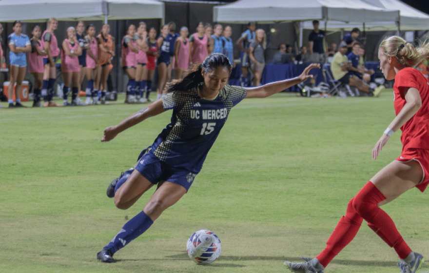 A woman in a blue UC Merced uniform prepares to kick the ball as a woman in a red uniform guards her