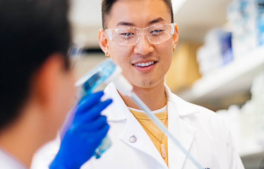 A researcher wearing a white lab coat and safety glasses smiles while examining a test tube 