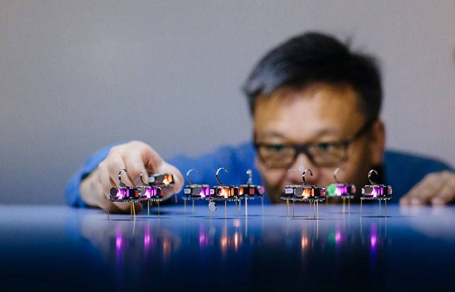 A man at table-level peers at tiny glowing robots on the table