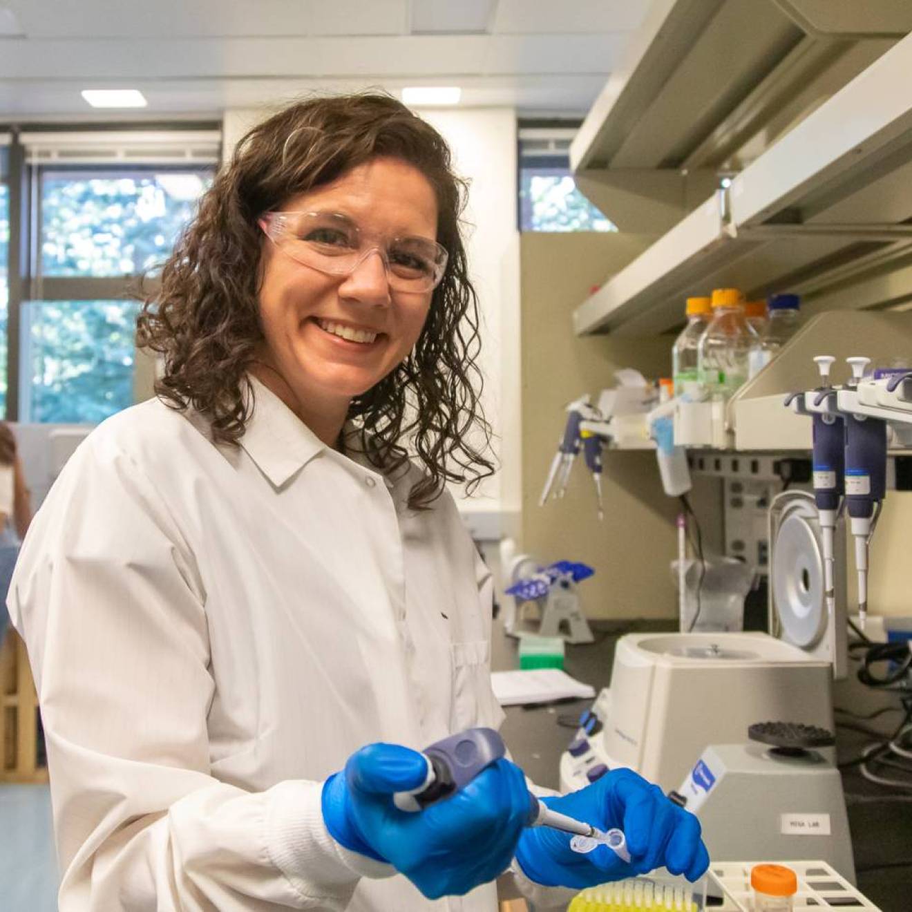 Karen Miga in safety glasses, white coat and blue gloves looks up from pipetting to smile at the camera in a lab