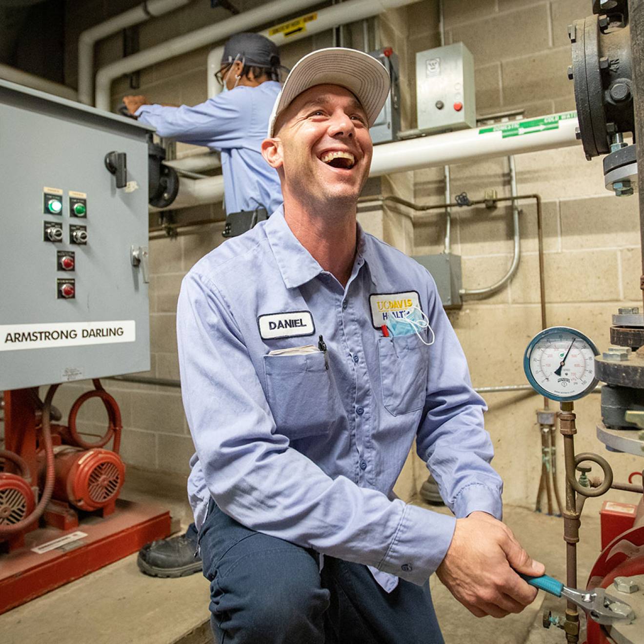 A maintenance worker in a light blue uniform kneels in a mechanical room, smiling as he uses a wrench to adjust a large pipe valve, while another worker operates a control panel in the background surrounded by pumps, gauges, and industrial piping.