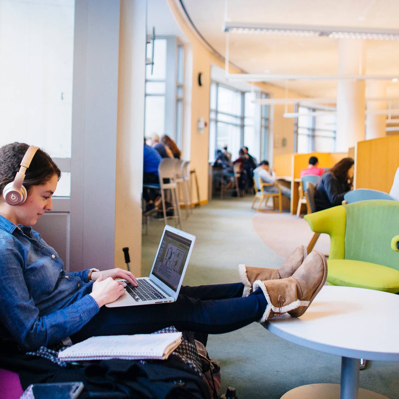 A woman sits on a sofa in a library typing on her laptop with her feet up in a table and headphones on.  Beyond her are more people studying at desks.