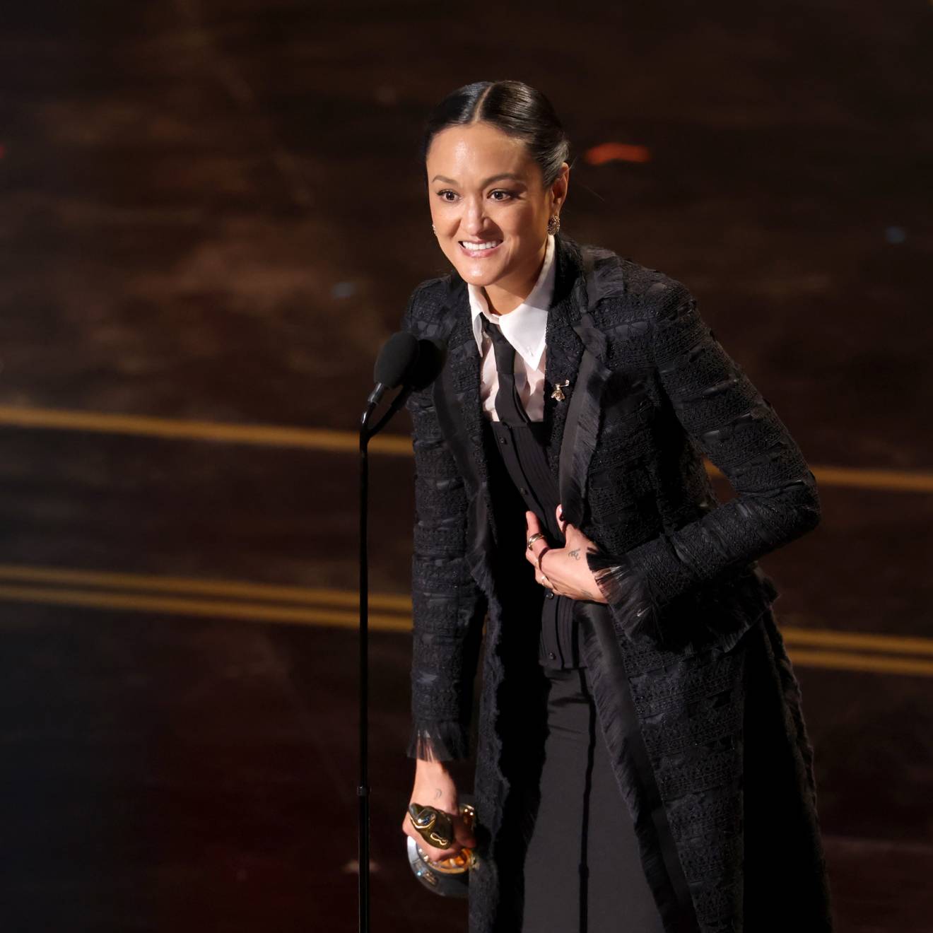 A woman speaks at a mic at the Oscars, holding the statuette and looking extremely happy