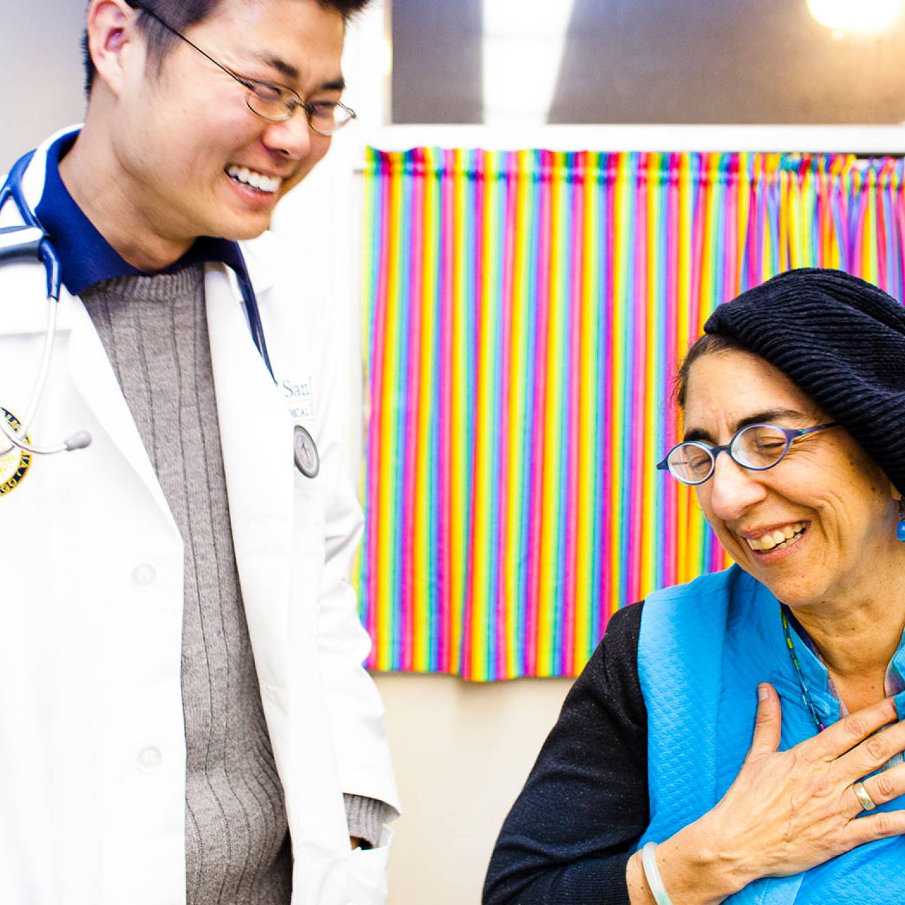 A doctor in a white coat laughs with a patient, who is smiling and holding her hand over her heart.