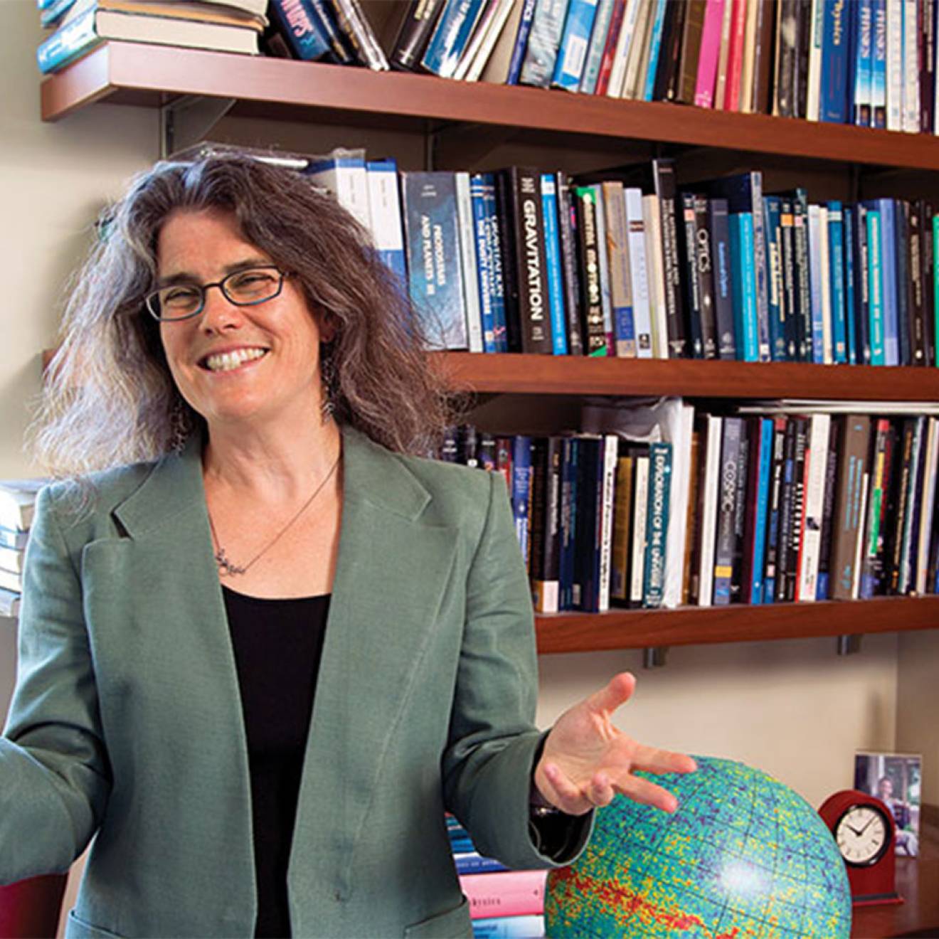 Andrea Ghez smiles and gesticulates with her hands, standing in front of a bookshelf and a globe