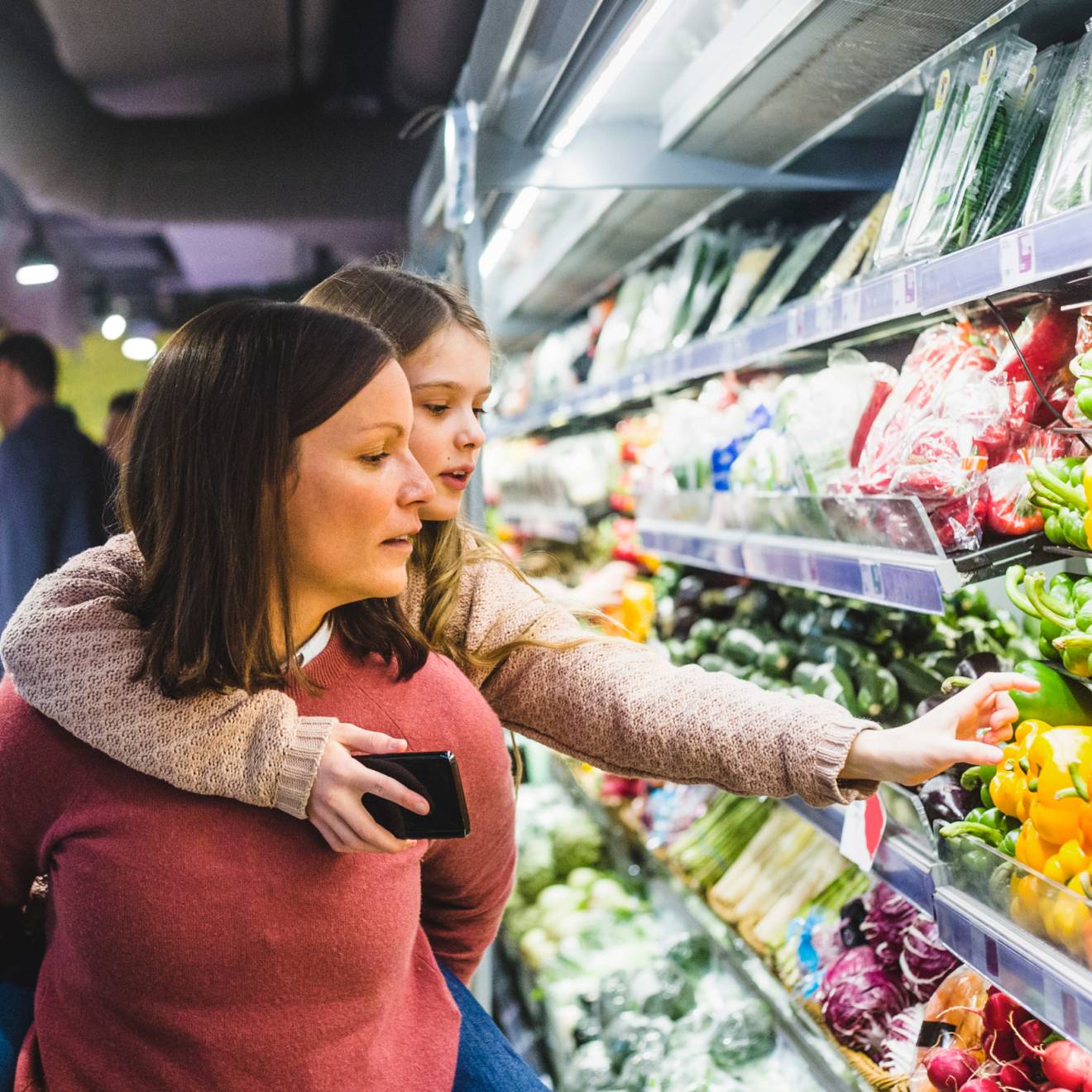 A child hanging on to her mom's back reaches for a pepper in the produce aisle