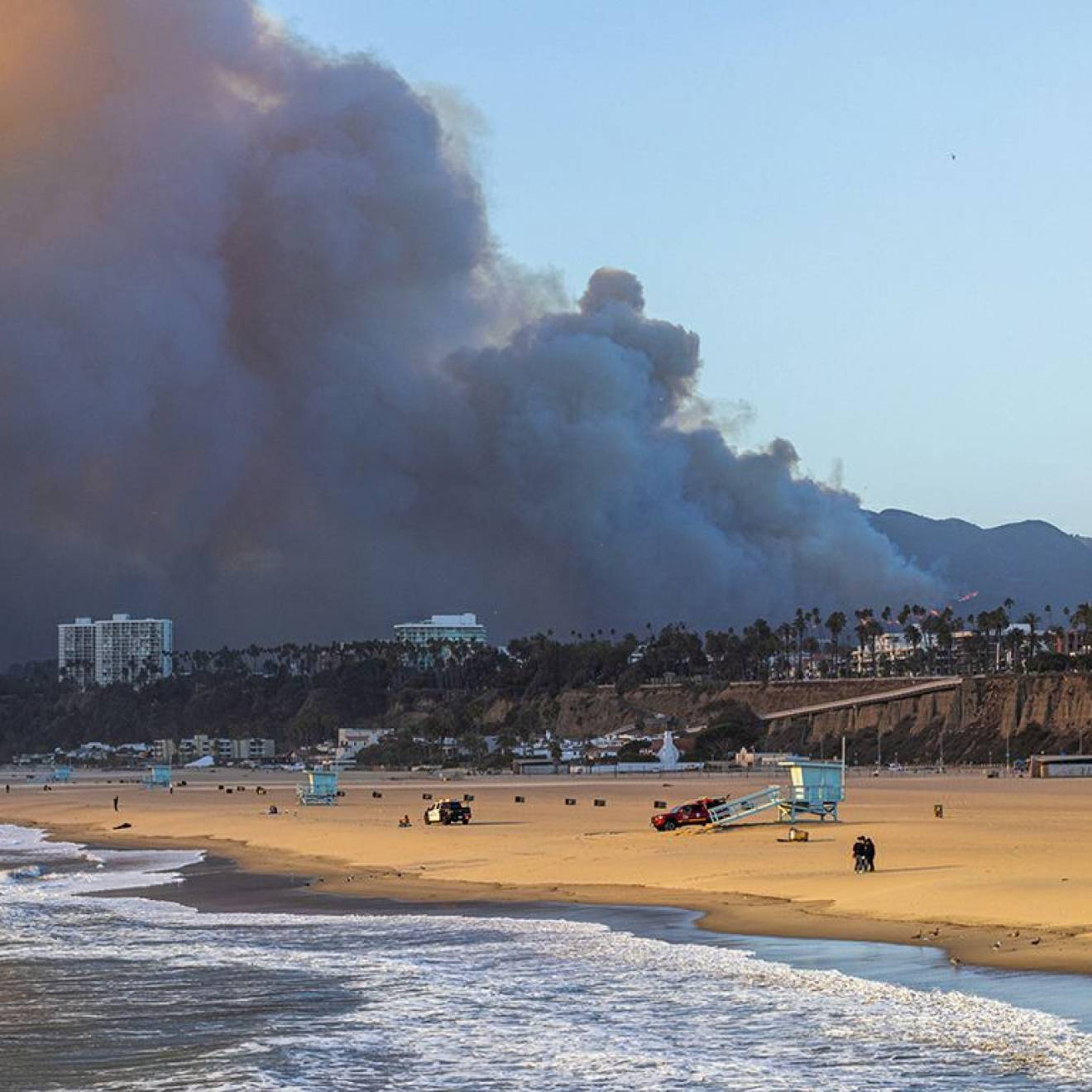 A wide sandy beach with gentle waves in the foreground, lifeguard towers and a few people scattered along the shore, while a massive dark plume of smoke rises from hills behind nearby coastal buildings under a clear blue sky.