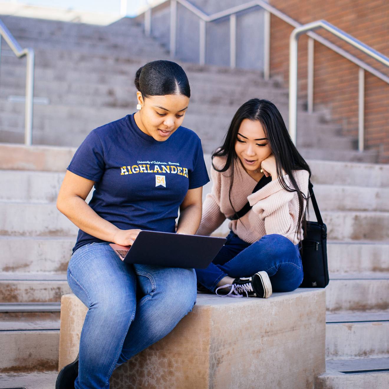 Two students sitting on a stairway landing outside looking at an open laptop