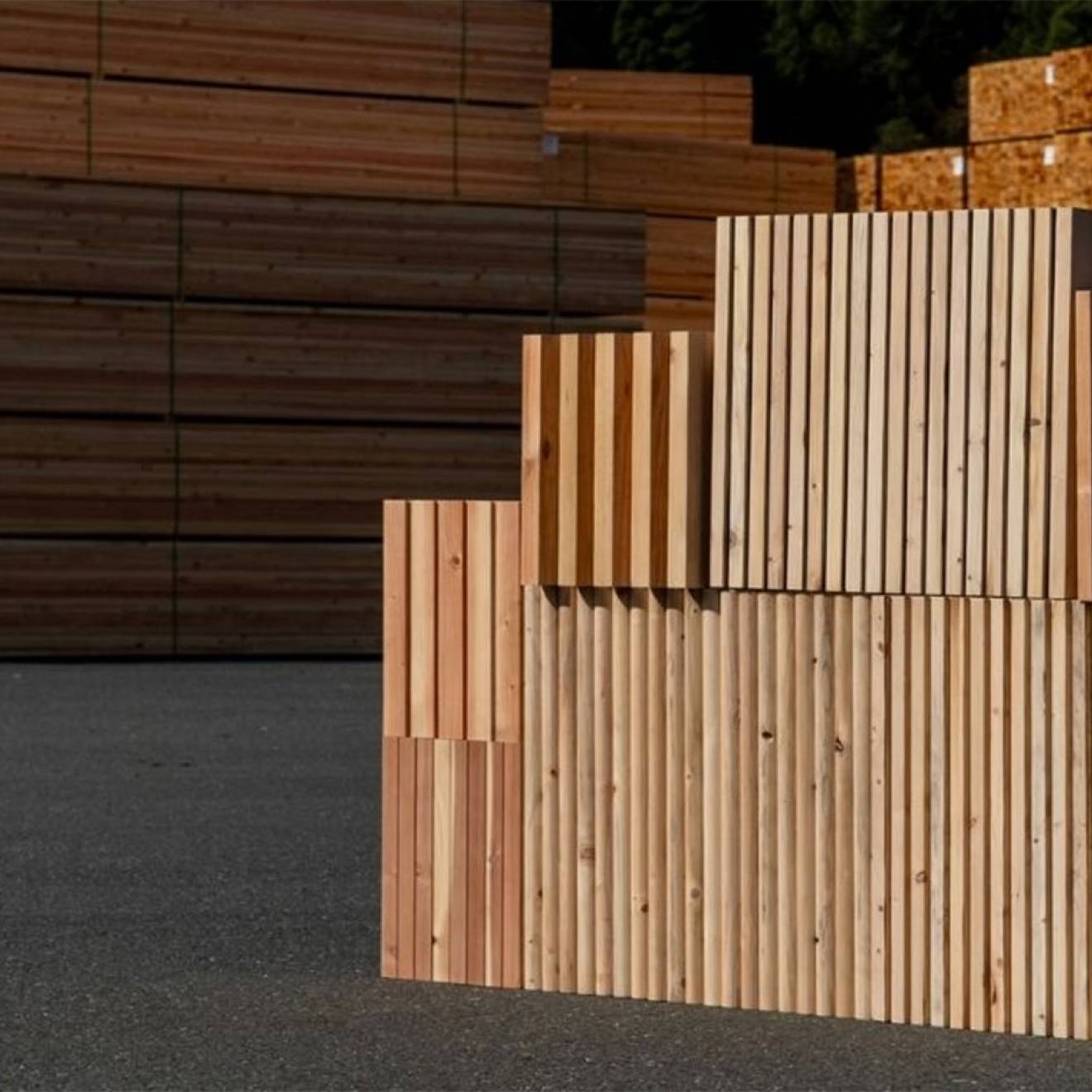 Stacked wooden panels with vertical slats arranged in stepped blocks on an outdoor asphalt surface, with large piles of lumber stacked in the background at what appears to be a timber yard.