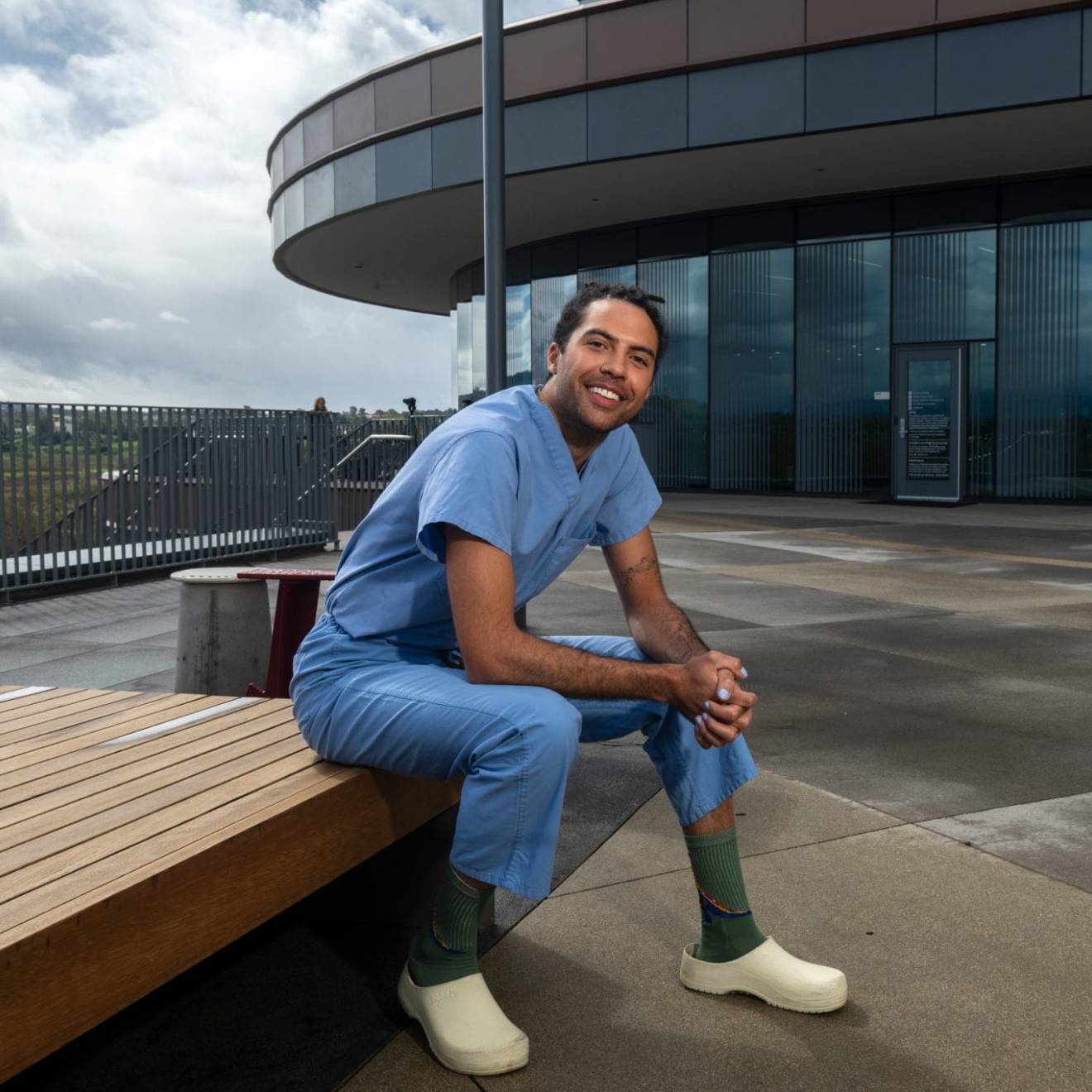 A person in blue scrubs sits outside a medical building and smiles