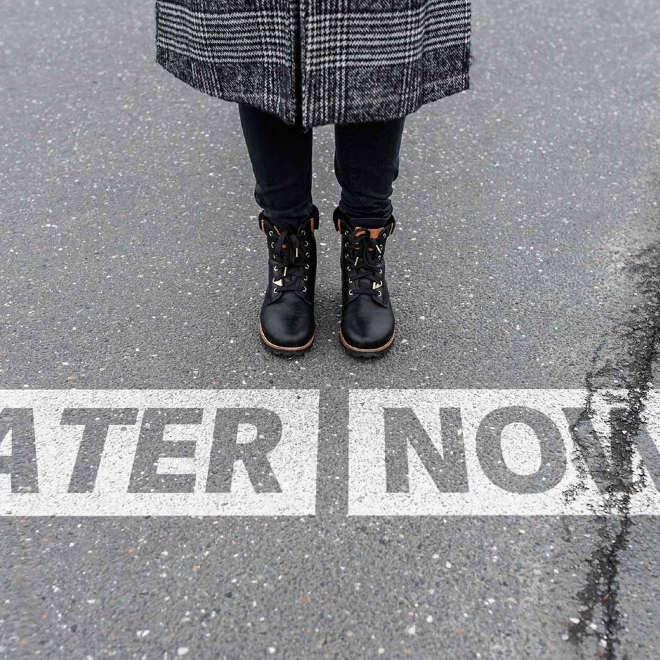 View of a person standing on pavement with only their legs and boots visible, positioned above two large white arrows painted on the ground—one pointing left labeled “LATER” and one pointing right labeled “NOW.”