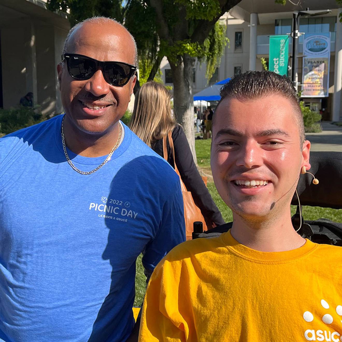 Ryan Manriquez, who uses a motorized wheelchair and wears a yellow shirt with the logo of the Associated Students of UC Davis on it, smiles for a photo alongside UC Davis Chancellor Gary May, who wears sunglasses and a blue t-shirt and is waving at the camera