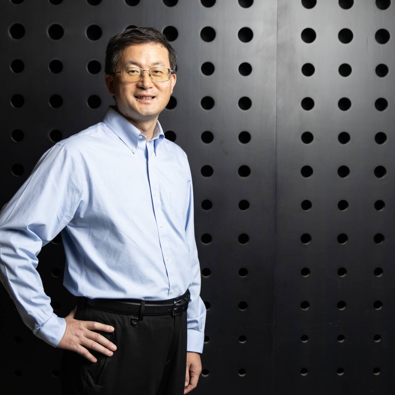 A man with glasses stands in front of a dark textured wall with his hand on his hip